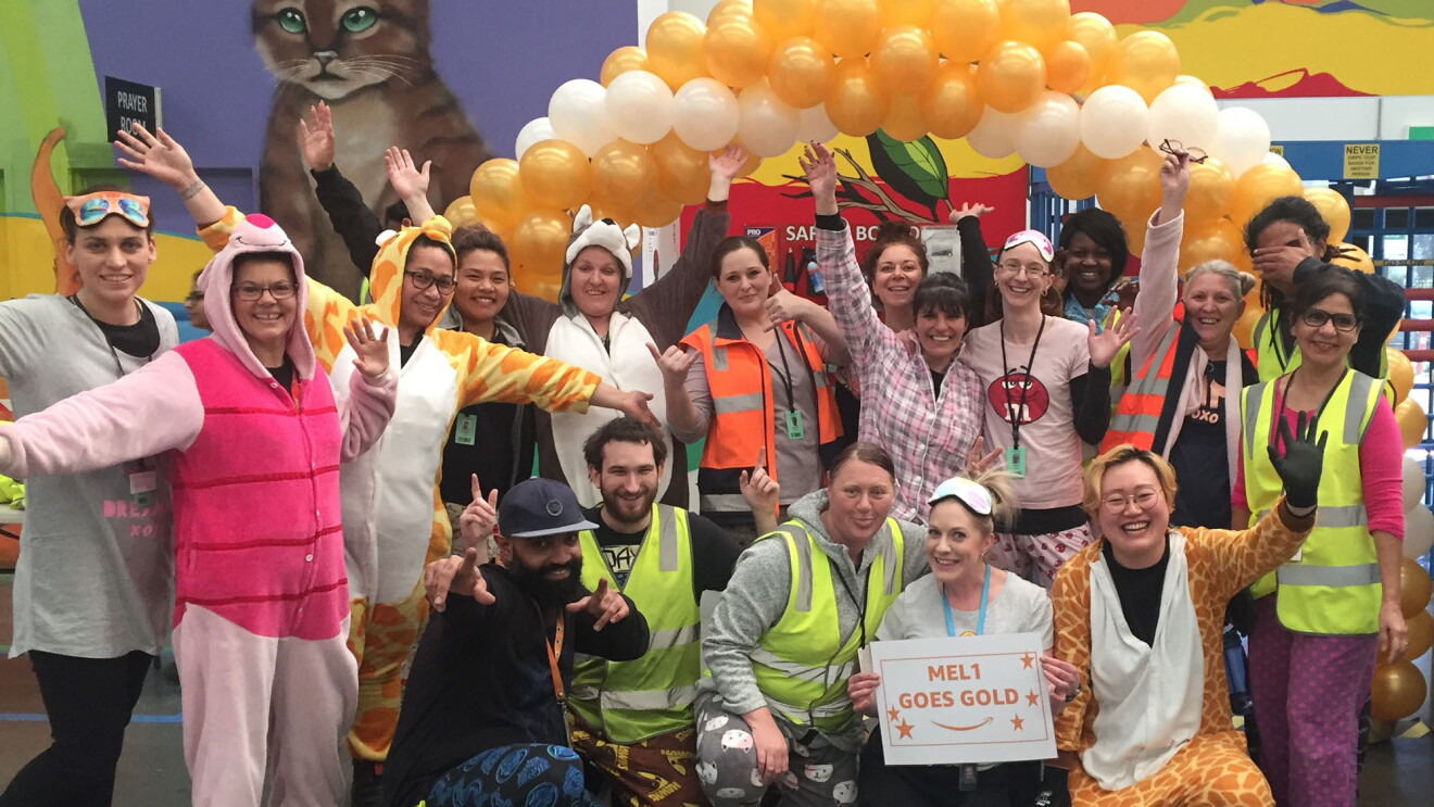 An image of Amazon employees wearing pajamas at work as a part of the Goes Gold campaign. They are standing in front of a balloon arch with gold and white balloons and a wall with a cat and a colorful background painted on it. 