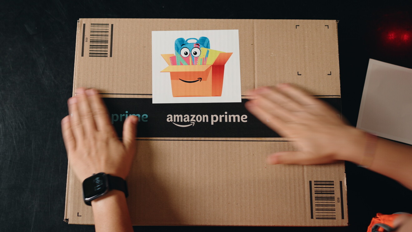 Una caja de Amazon de color marrón sellada con la cita negra. En el centro de la caja, una etiqueta que es una caja de la que sale material escolar con una mochila, libretas y bolis. Hay dos manos que intentan abrir la caja.