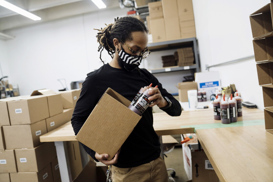 An employee of Capital City Mambo Sauce wears a face mask while filling a box with Capital City Mambo Sauce. Behind the employee are stacks of boxes.