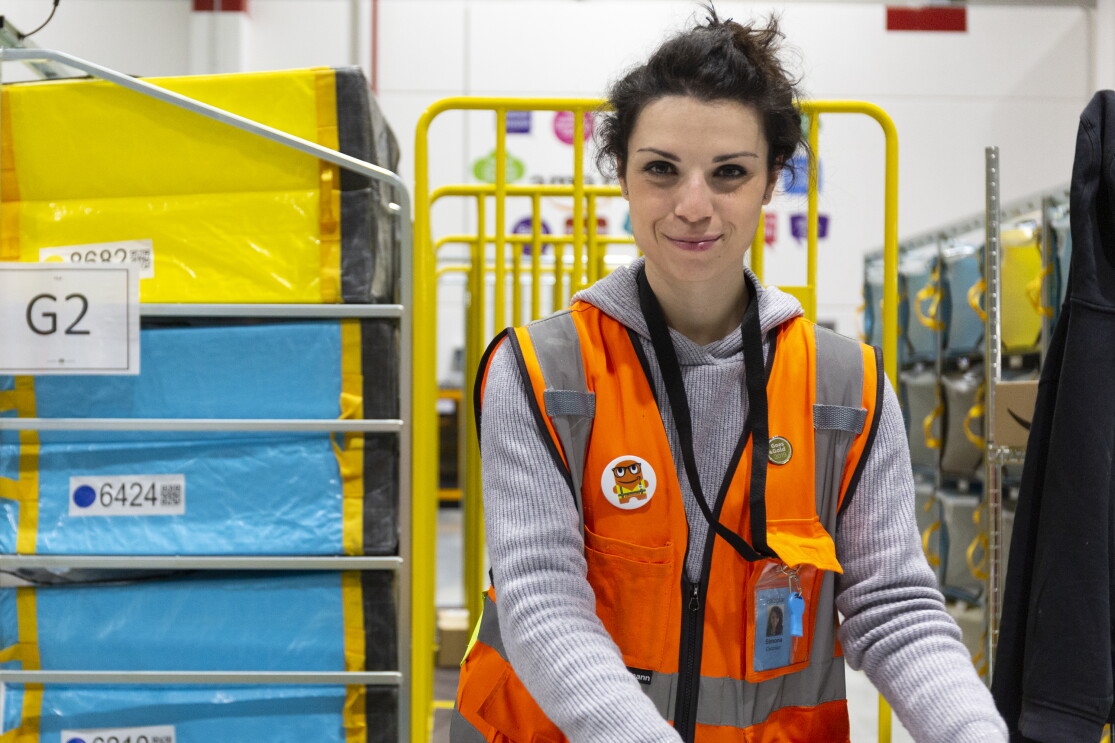 Simona Calzolari, operatrice di magazzino, inquadrata nel deposito di smistamento di Brandizzo. Indossa un giubbotto arancione ed è circondata da carrelli gialli e pacchi celesti e gialli. 