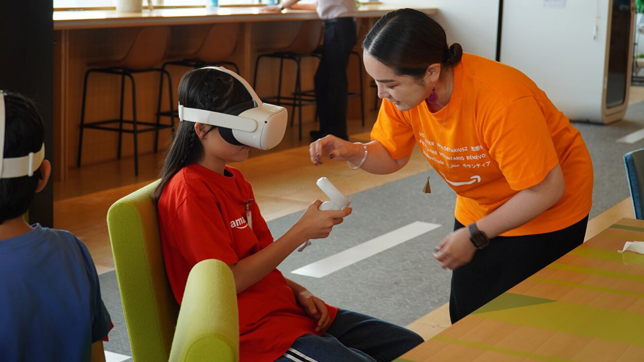 広い室内で、白いVRゴーグルを顔に付けて赤いTシャツを着た児童が椅子に座っている。手には白いコントローラーを持っている。右側にオレンジ色のTシャツを着た女性がいて、児童をサポートしている