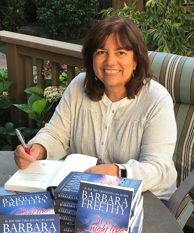 Amazon author Barbara Freethy sits on a deck in the sunshine behind a stack of her books. She is autographing one book.