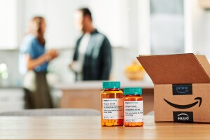 Two bottles of medication are on a table with an Amazon box next to them. The background is out of focus and two adults talk in a kitchen.