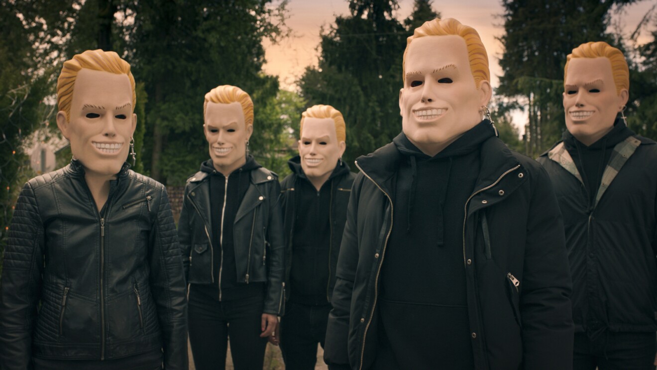 Five figures in black jackets wearing identical blonde masks with wide grins