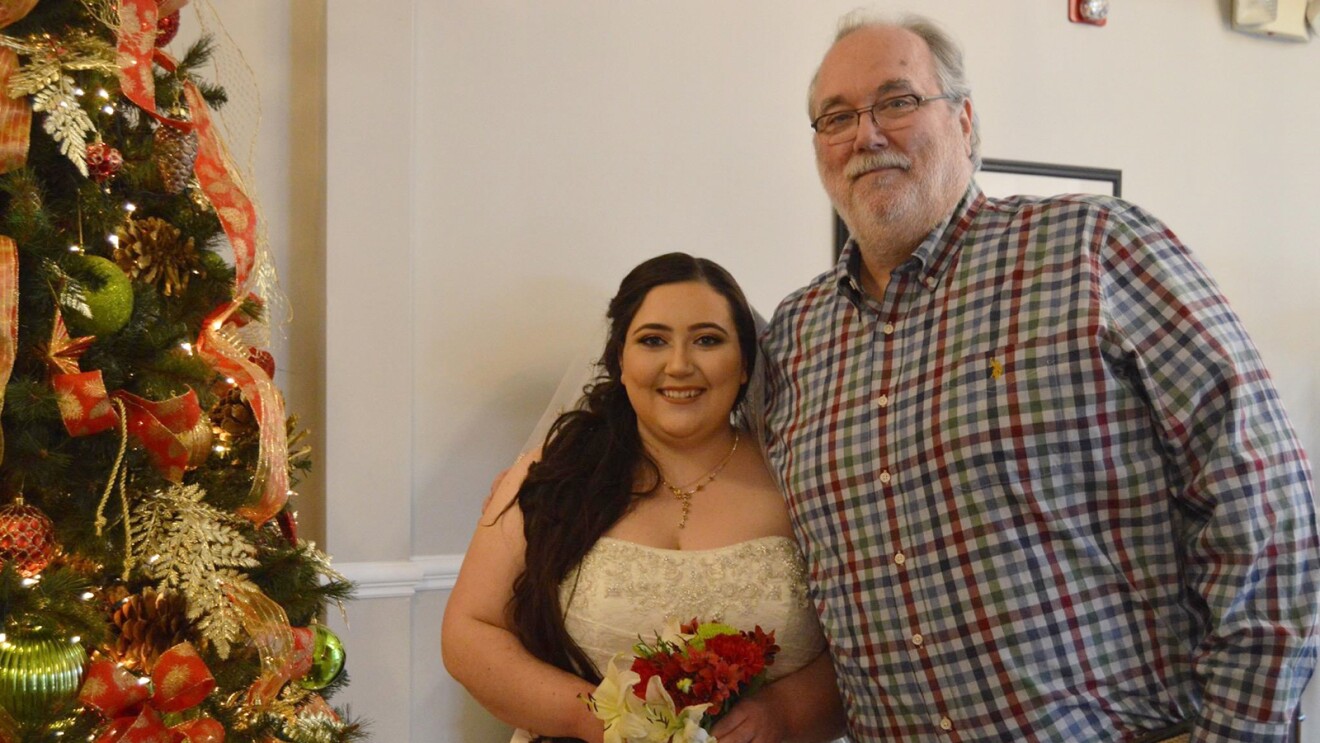 An image of a man and his adult daughter standing for a photo at her wedding. There is a Christmas tree beside them and the daughter is wearing a wedding dress and veil while holding a small bouquet of roses.