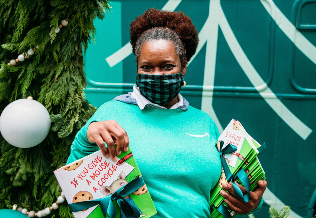 An image of a woman smiling for a photo while wearing a mask. She is holding stack of children's books and wearing a green Amazon shirt. Behind her is a large wreath with ornaments on it.