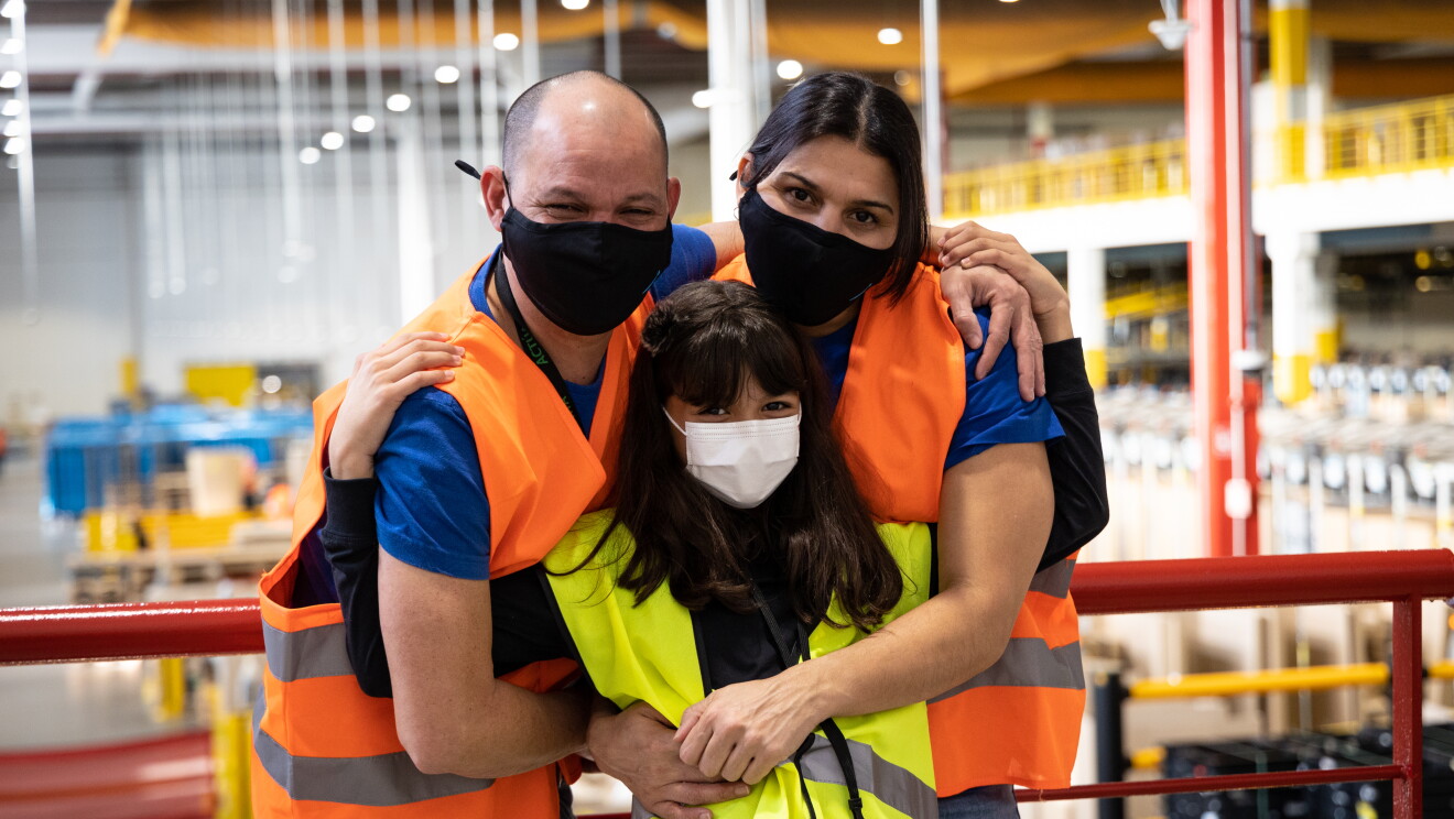 En unas escaleras del centro logístico de San Fernando de Henares está Pepe y Laura con su hija de pie apoyados en una barandilla de color rojo. Los tres miran a cámara y su hija está en el medio. Pepe y Laura van con jeans, camiseta azul de manga corta y un chaleco de color naranja. Llevan una mascarilla negra. Pepe lleva la cabeza rapado y Laura tiene el pelo  negro y lo lleva recogido. La niña tiene el pelo largo, lleva una camiseta de manga larga negra y un chaleco de color amarillo. 