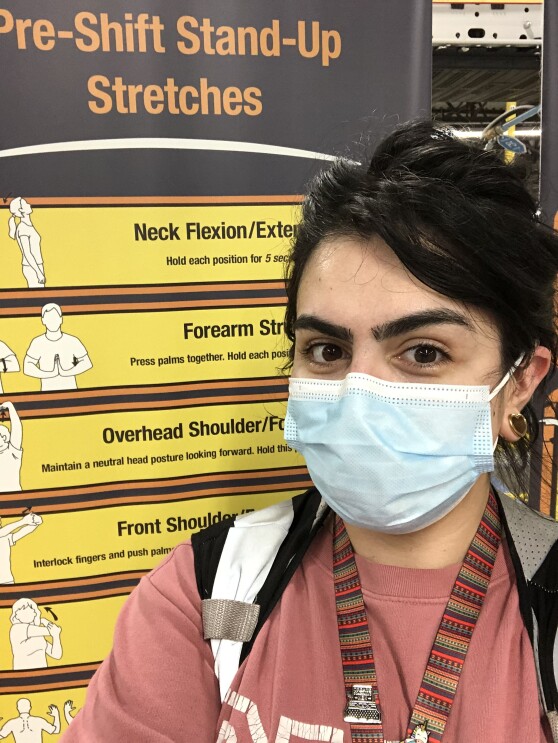 Beka Perez, an Amazon associate, smiles for a photo in front of a sign at an Amazon fulfillment center. The sign shows useful stretches for fulfillment center employees. Beka is wearing a face mask. 