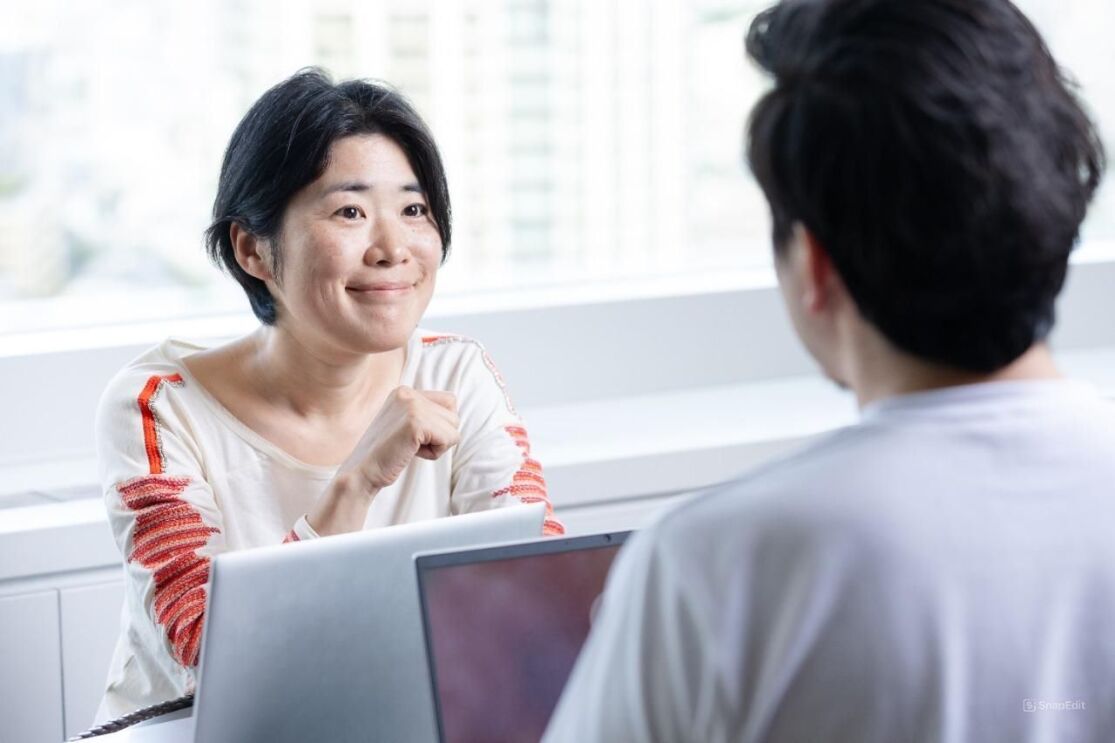 白いシャツを着ている女性が男性1人と向き合って座っている。彼らの前にはそれぞれパソコンが置かれている。女性の後ろの窓からは明るい外の風景が見える