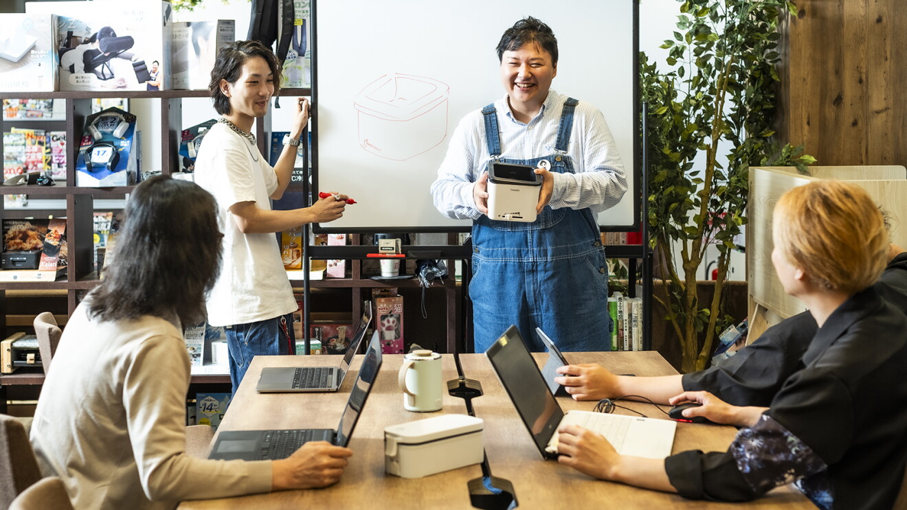 商品を手にホワイトボードの前で社員たちに話しかける男性（﨏さん）