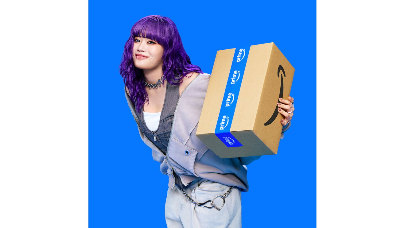紫色の髪の毛の若い女性が、左手にAmazonロゴ入りダンボール箱を抱えてポーズを取っている。