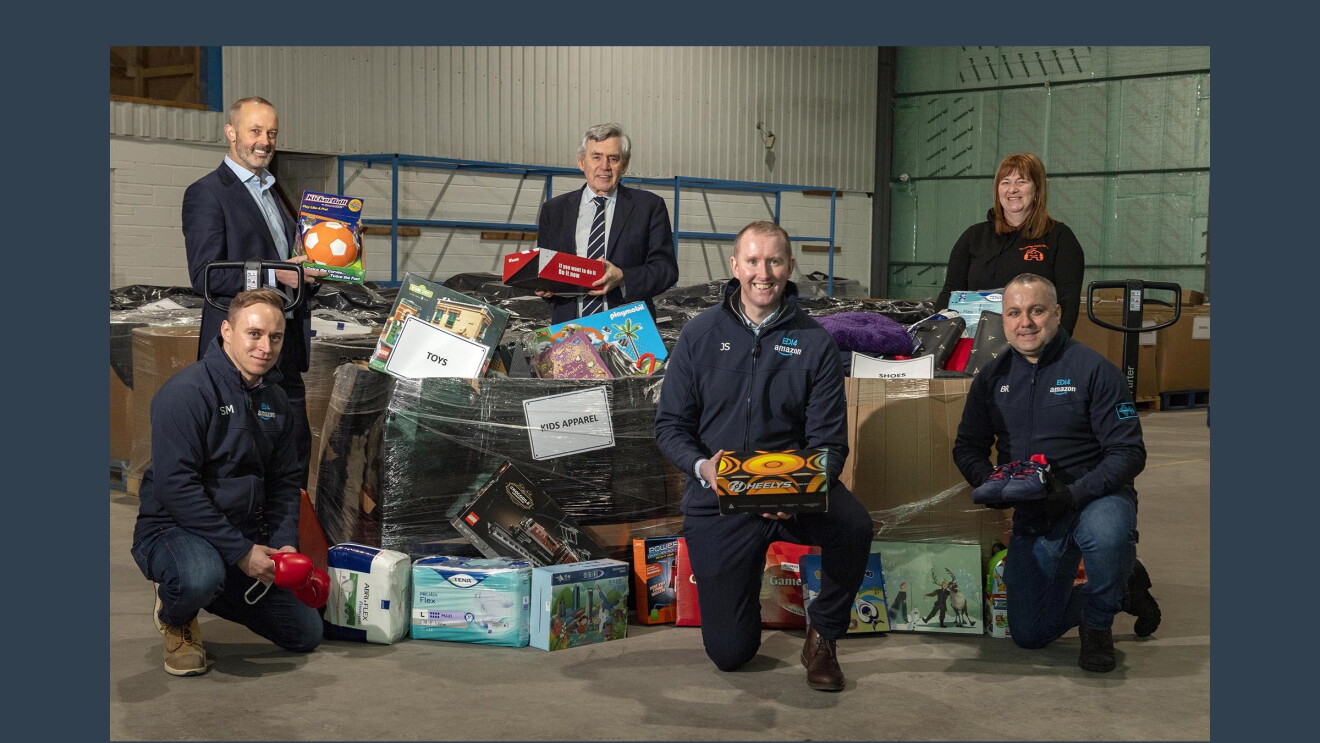 Six individuals pose together in a warehouse setting with products to be donated to those in need