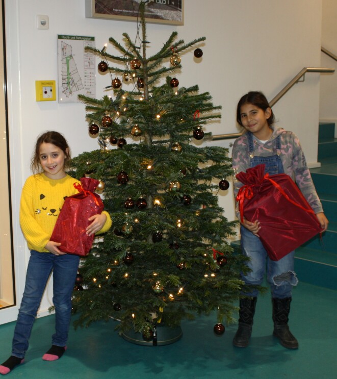 Zwei Kinder halten Geschenke in den Armen und stehen neben einem Weihnachtsbaum.