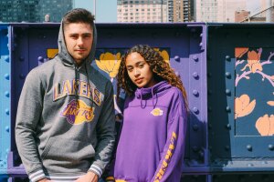 An image of a man and a woman standing outside in front of a metal wall with colorful, yellow, purple, and blue murals painted on it. The man is wearing a gray hoodie that says "Lakers" on it with a Lakers logo, and the woman is wearing a purple hoodie with the Lakers logo on the left chest and yellow text that says "Lakers" on the sleeve.