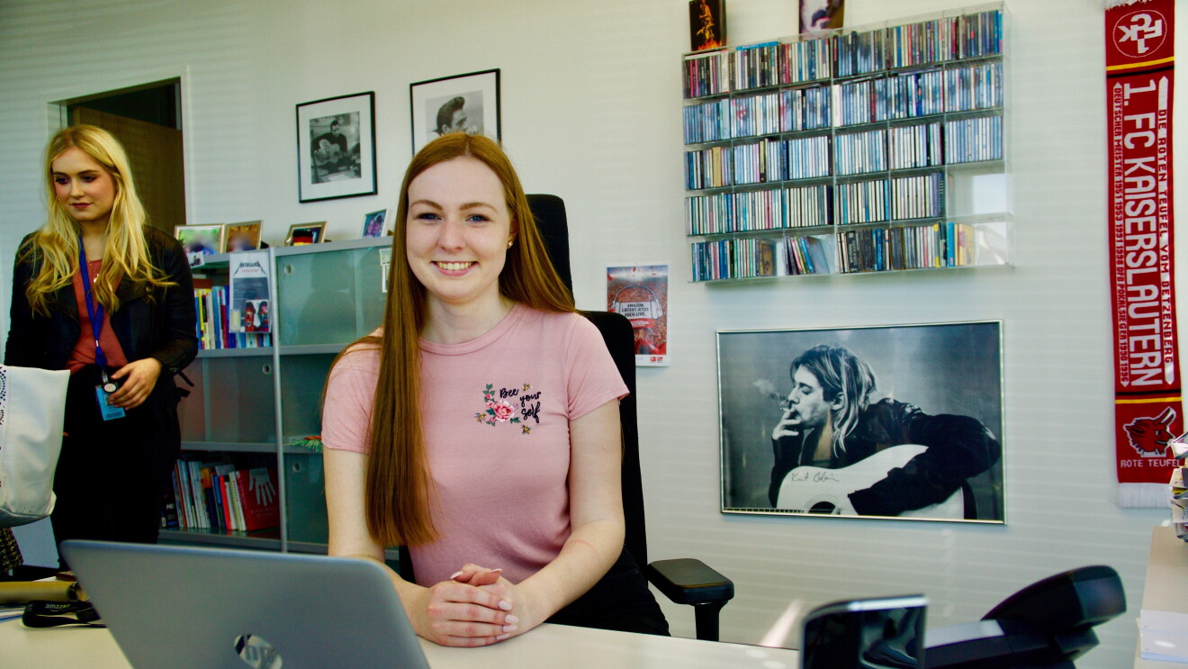 Eine junge Frau in rosa T-Shirt sitzt an einem Schreibtisch, vor ihr ein Laptop