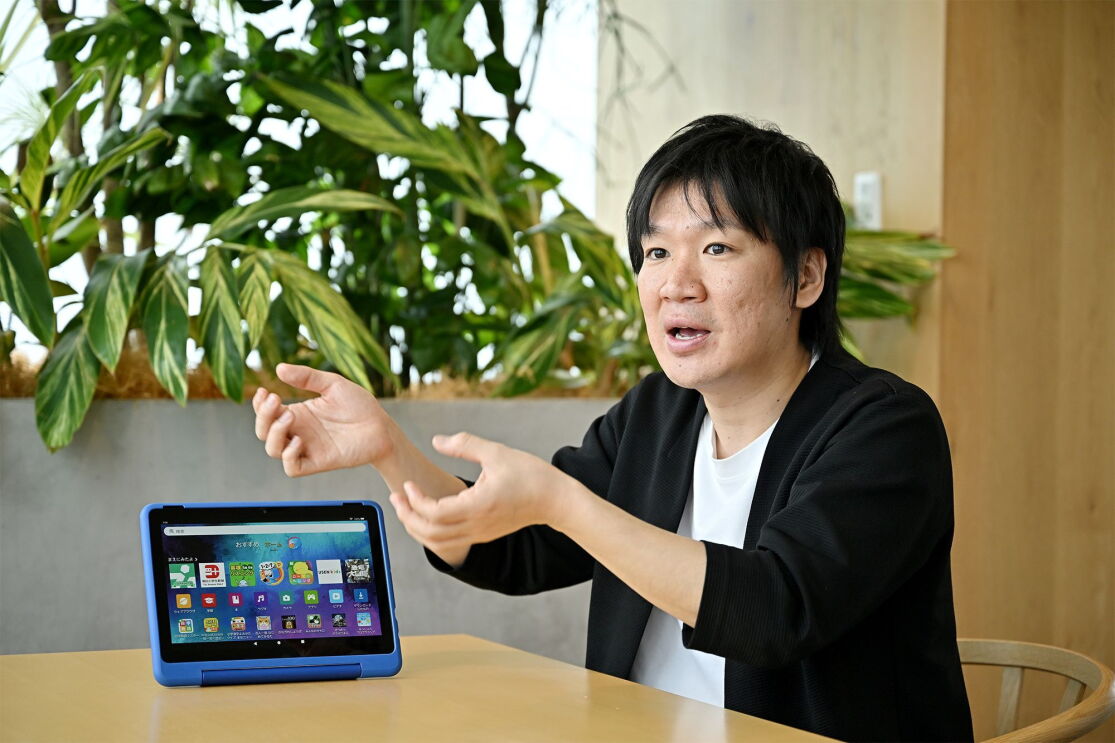 白いシャツに黒いジャケットを着た男性が身振り手振りで話している。テーブルに青いカバーがついたタブレットが置かれている。