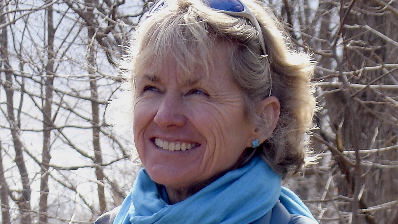 Amazon author Kristiana Gregory, a woman with blond hair, smiles looking to the left. She is wearing a blue scarf and blue square earrings. It's winter, and trees with no leaves are in the background. She has sunglasses on her head. 