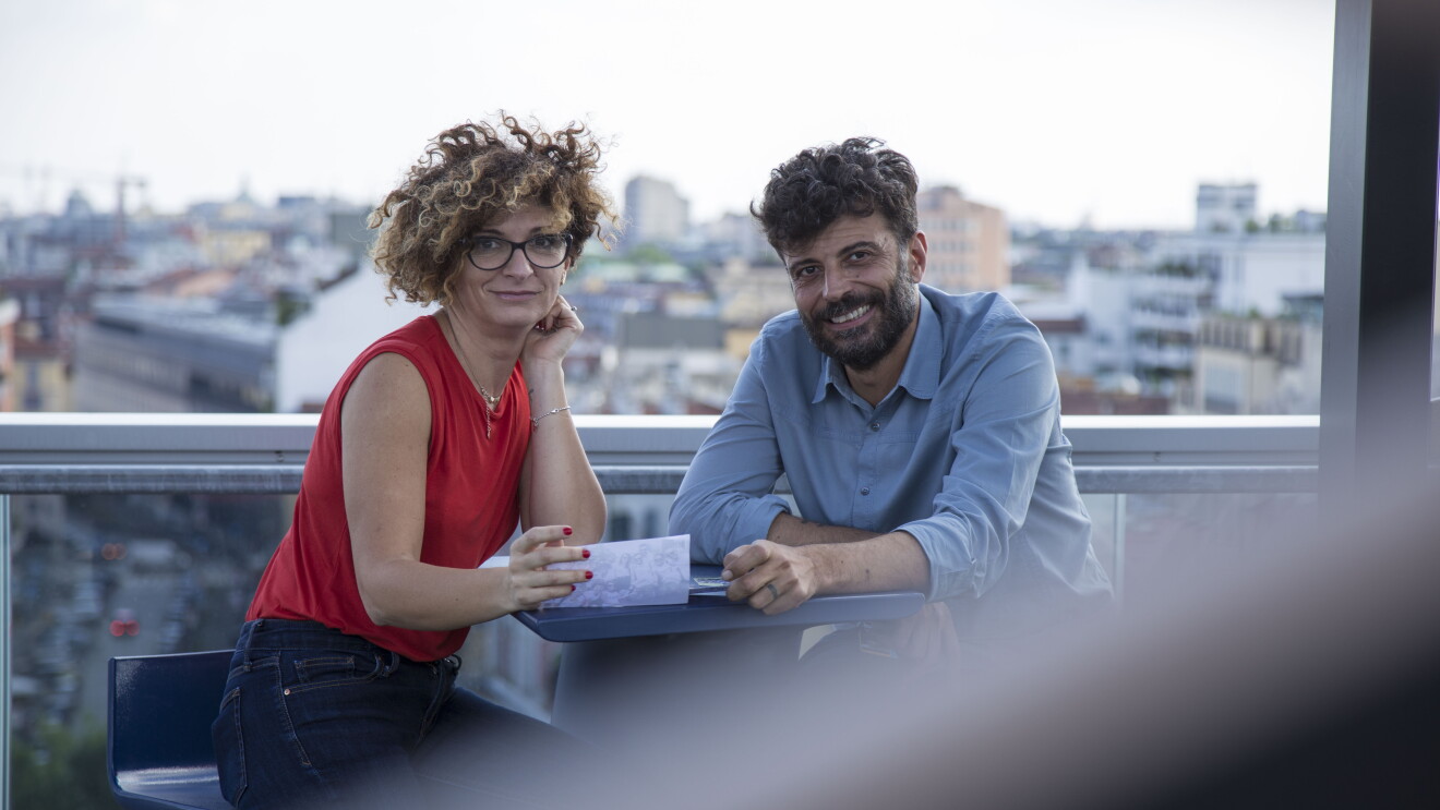 Due persone siedono allo stesso tavolo, sulla terrazza del palazzo Amazon di Milano. A destra, Alessandro Saccon, Consumer PR Manager. Di fronte a lui siede Alessandra Meucci, Executive Assistant. Sullo sfondo lo skyline di Milano. La foto è stata scattata in occasione del Pride di Milano di giugno 2018. 