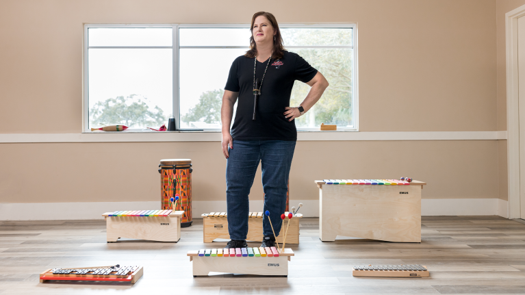 A photo of Nicole Greggs, president and owner of MusicMakers! Greggs is standing in a classroom with a variety of xylophones, drums, and other musical instruments.