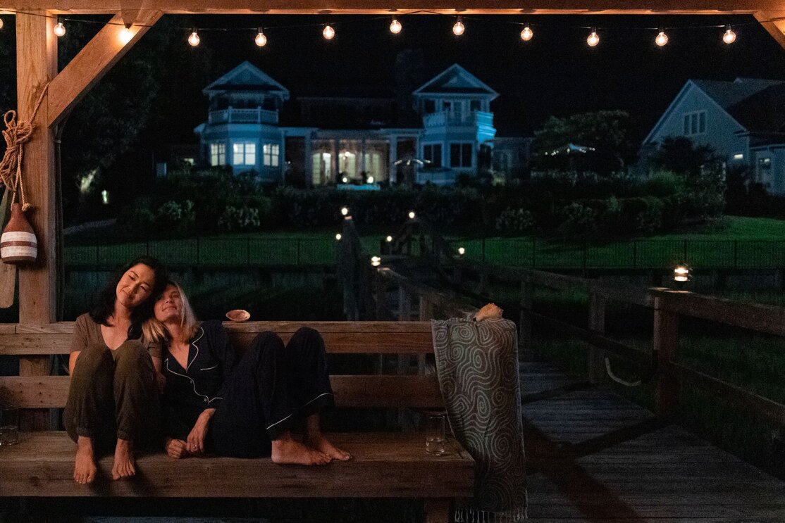 An image of two friends sitting close to each other on a bench on a boat dock at night. One of the women has her head on the other's shoulder and they're smiling and looking off into the distance.
