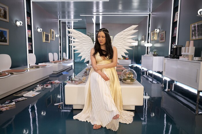 An image of Constance Wu sitting on a table in a clinical waiting room. She is wearing a long, white dress, angel wings, and a small, gold crown.