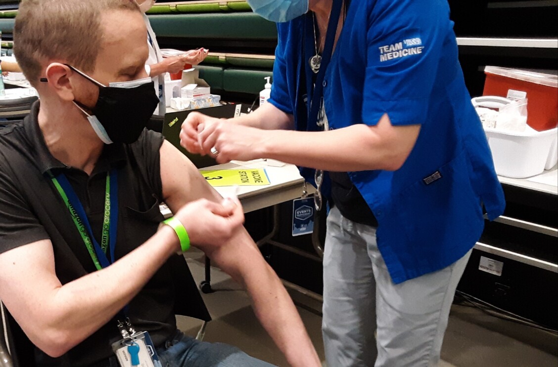 A man rolls up his sleeve while a medical professional administers the COVID-19 vaccine in his arm. Bot individuals are wearing masks.