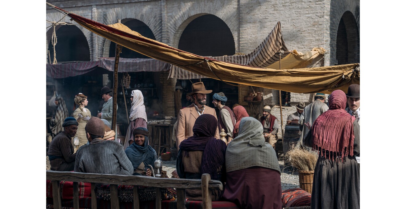 中東の屋外マーケットのような場所を歩く帽子の男性。周囲には布を巻いている人が多いが、西洋の服装をした人物も見受けられる。人々の情報に布の幕が張られている。