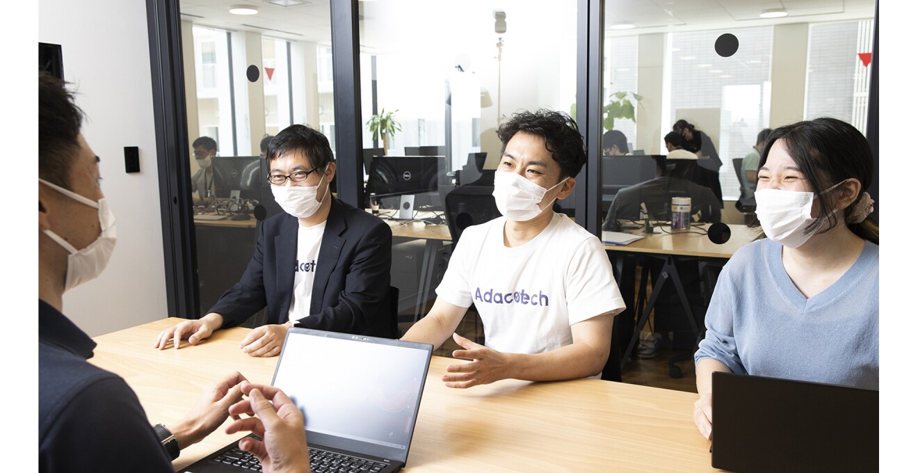 社内ミーティングの写真。ガラスのパーテーションの向こうにたくさんの社員がいるのが見える。