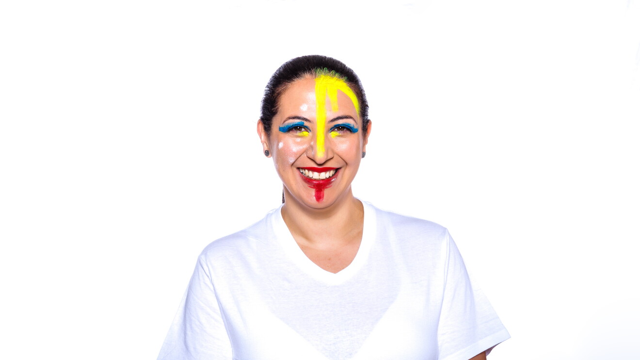 Para el Día de la diversida Amazon ha preparado una acción con asociados de diferentes países que llevan la bandera pitnada en su cara. En la foto, Giselle con camiseta blanca, mirando sonriendo a cámara con la bandera de su país puntada en la cara. Tiene el pelo negro oscuro recogido con una coleta detrás. Lleva pendientes en las orejas. 