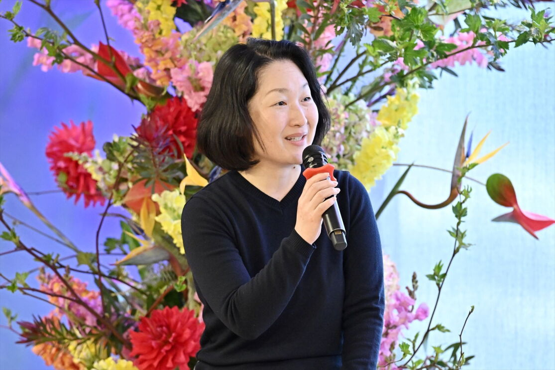 黒いシャツを着ているショートヘアの女性が大きな花の装飾の前でマイクを持って話している。