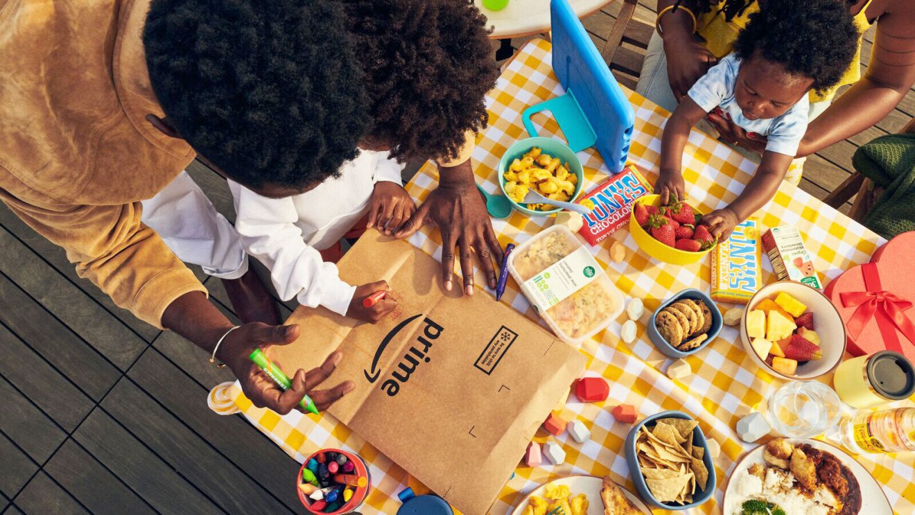 Ein Bild von einem Tisch voller Essen. Am Tisch sitzen zwei Kinder mit ihren Eltern. Ein Elternteil hilft einem Kind beim Ausmalen einer Amazonas-Box.