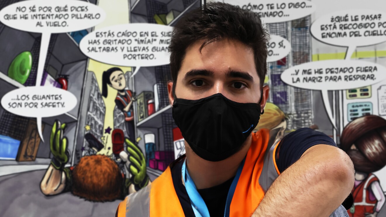 Santiago Morilla es empleado en el centro logístico de Illescas en Toledo.  Santiago Morilla delante de sus ilustraciones en forma de cómic gigante. Santiago está sentado delante de una de sus viñetas. Él tiene el pelo negro y corto. Va con una camiseta azul de algodón de manga corta y un chaleco de color naranja con el badge en el cuello. Lleva unos jeans y tiene la mano izquierda apoyada en su pierna izquierda, que está doblada, y la mano derecha apoyada en la pierna derecha. De fondo las viñetas gigantes. Va con una mascarilla de color negro.