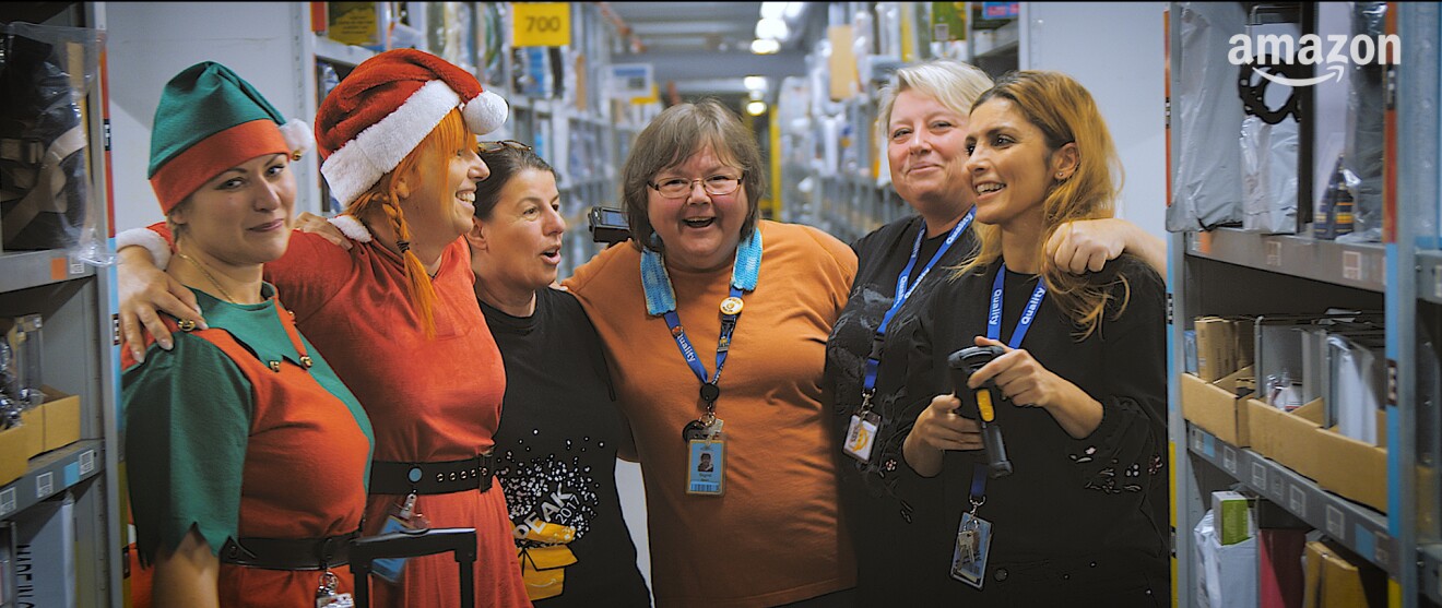 EineGruppe von 6 Mitarbeiterinnen steht in einem Regalgang. Eine trägt eine Weinachtszipfelmütze, ein andere ein Elfenkostüm.