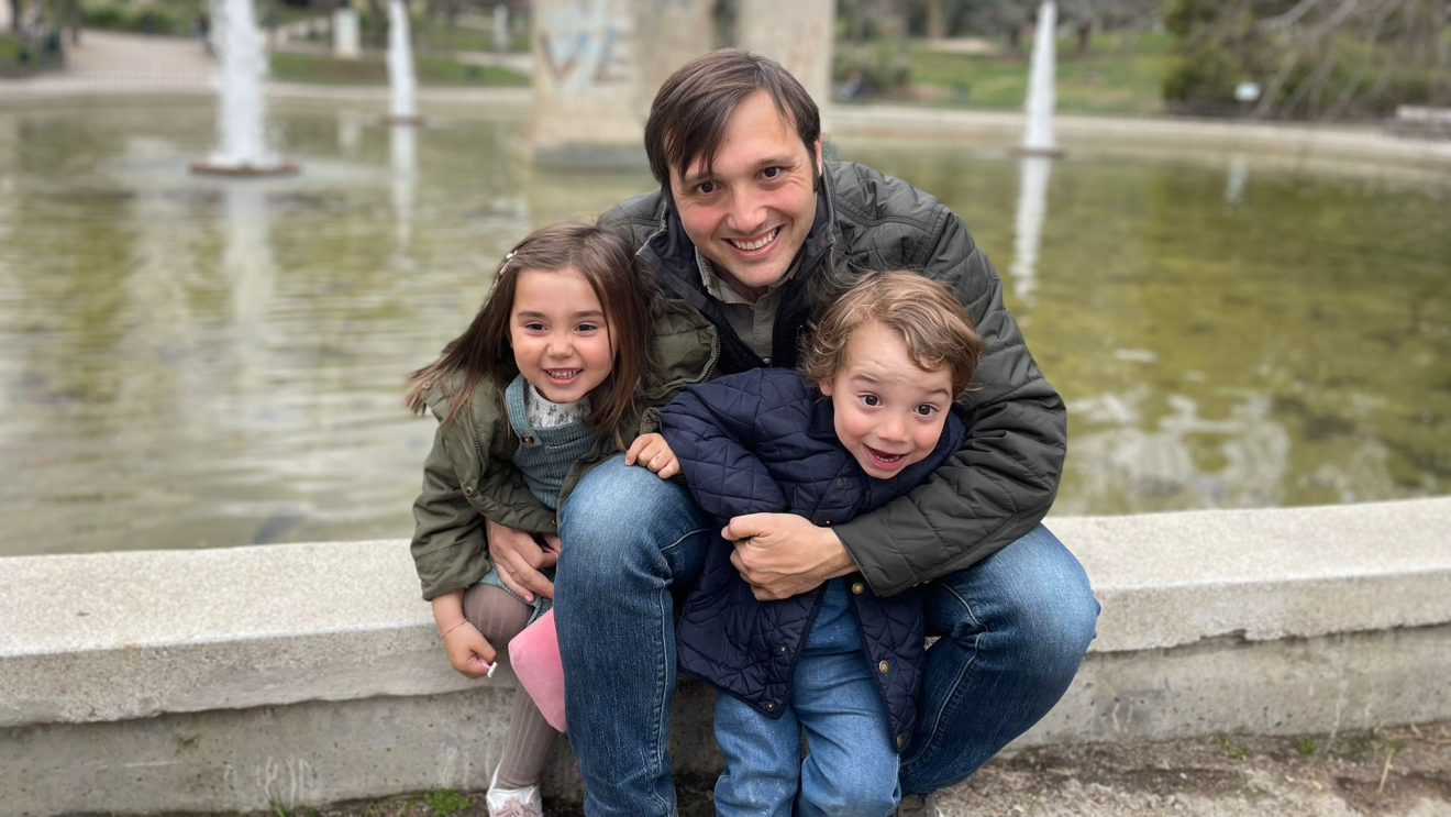 Alberto Saavedra con su hija y su hijo en el parque de Berlín, delante de un estanque con unas piedras de decoración dentro. Los tres están sentados apoyados justo delante del estanque. Alberto va con una chaqueta marrón, tiene el pelo corto con plequillo y mira a cámara sonriendo. La hija tiene el pelo largo, va con una chaqueta verde, un peto y medias y deportivas. Su padre le agarra con el brazo derecho. Con el brazo izquierdo agarra a su hijo con unos jeans y un chaqueta azul. De fondo el estanque y árboles en invierno, algunos con hojas y otros sin hojas.  