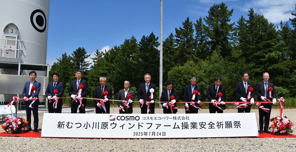 青空の下、赤いカーペットの上で11人のスーツを着た男性が紅白のテープカップをしている。その手前に新むつ小川原ウィンドファーム操業安全祈願祭という大きな白いパネル