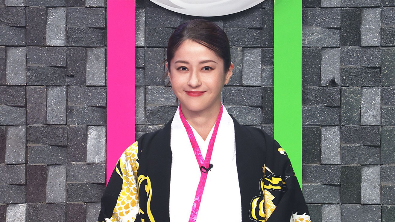 灰色のれんがを組み合わせたような壁を背景に、白の着物に金色の模様入り黒い羽織を身に着けた女性の上半身。壁にはピンクと黄緑の装飾帯が縦に配置されている。