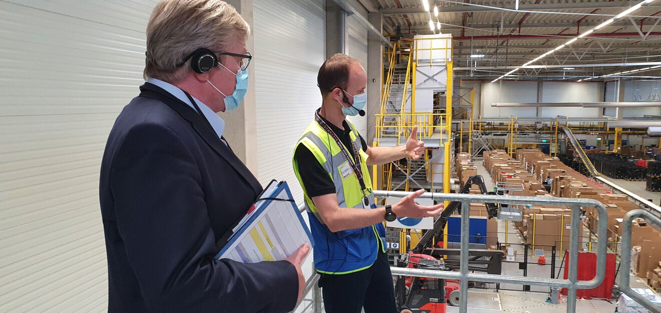 Ein Herr im blauen Anzug (links() mit einem Amazon Mitarbeiter in Sicherheitsweste: Beide stehen auf einer Ballustrade und blicken von oben in die Halle.