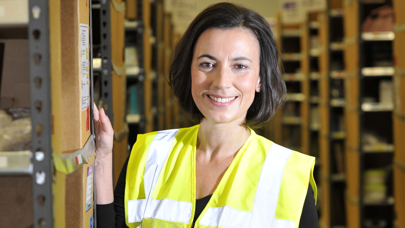 Eine Frau mit halblangem Pagenschnitt und braunen Haaren lehnt leicht an ein Regal eines Logistikzentrums. Sie trägt eine gelb-blaue Sicherheitsweste.