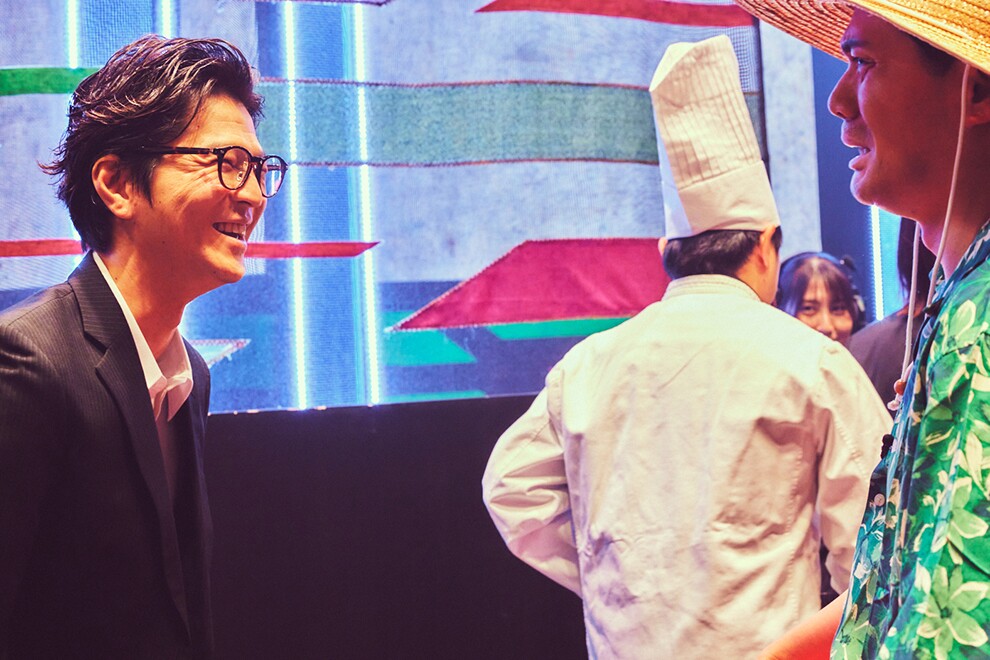 派手な柄の垂れ幕を背景に立つ2人の男性。左側の男性はスーツと眼鏡で笑顔、向かい合う右側の男性は緑のアロハシャツと麦わら帽。奥側に白いコック姿の人物の後ろ姿。
