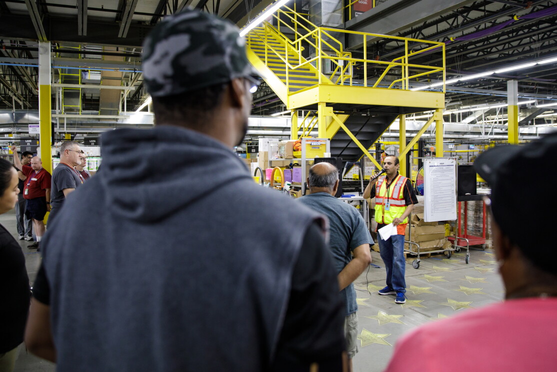 People stand in a warehouse space and look toward a man in a yellow safety vest who is speaking into a microphone.