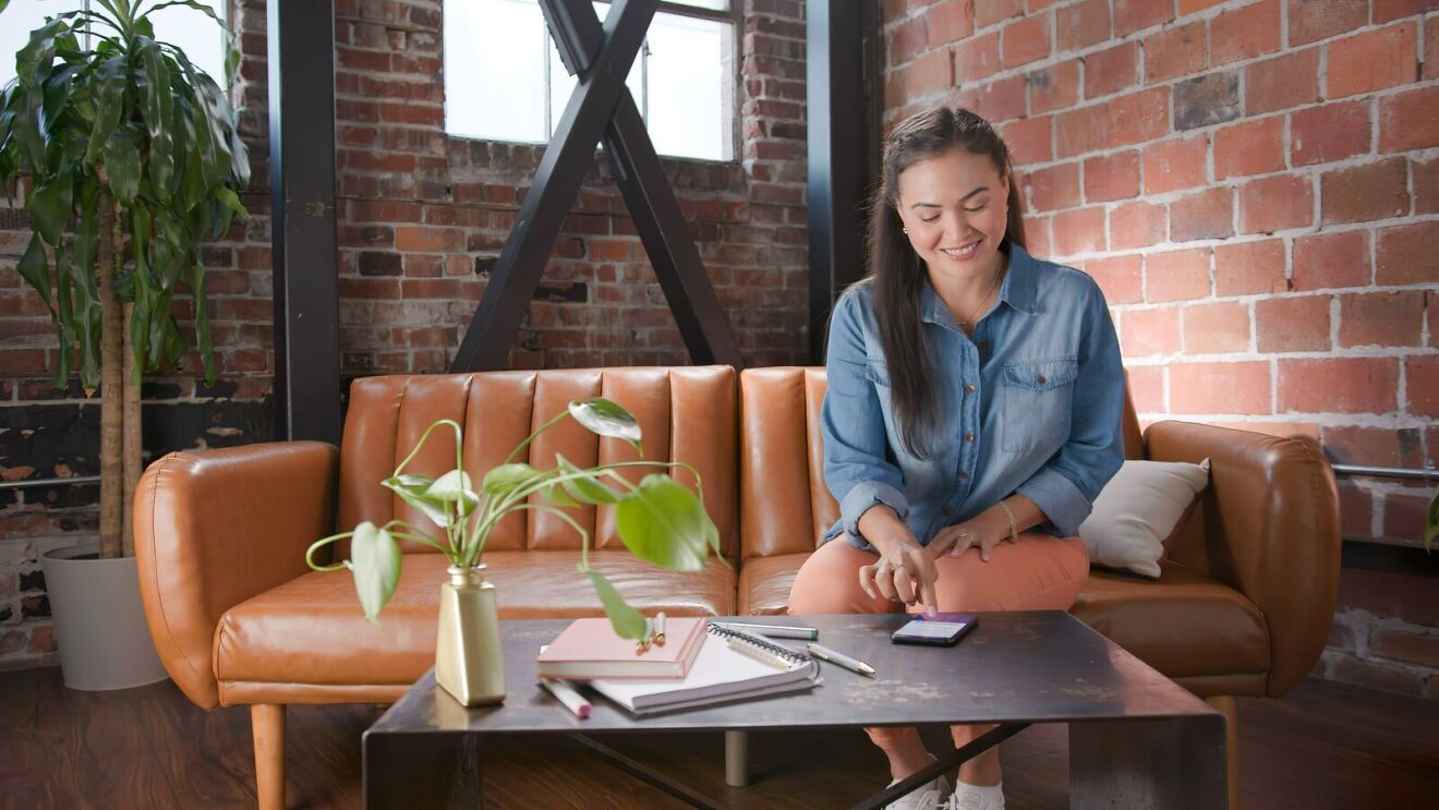 An image showing a woman seated on a leather sofa using her phone which is on the table.