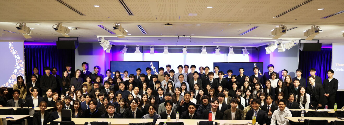 広い会議室で集合している150人以上の人々。多くはスーツを着ている