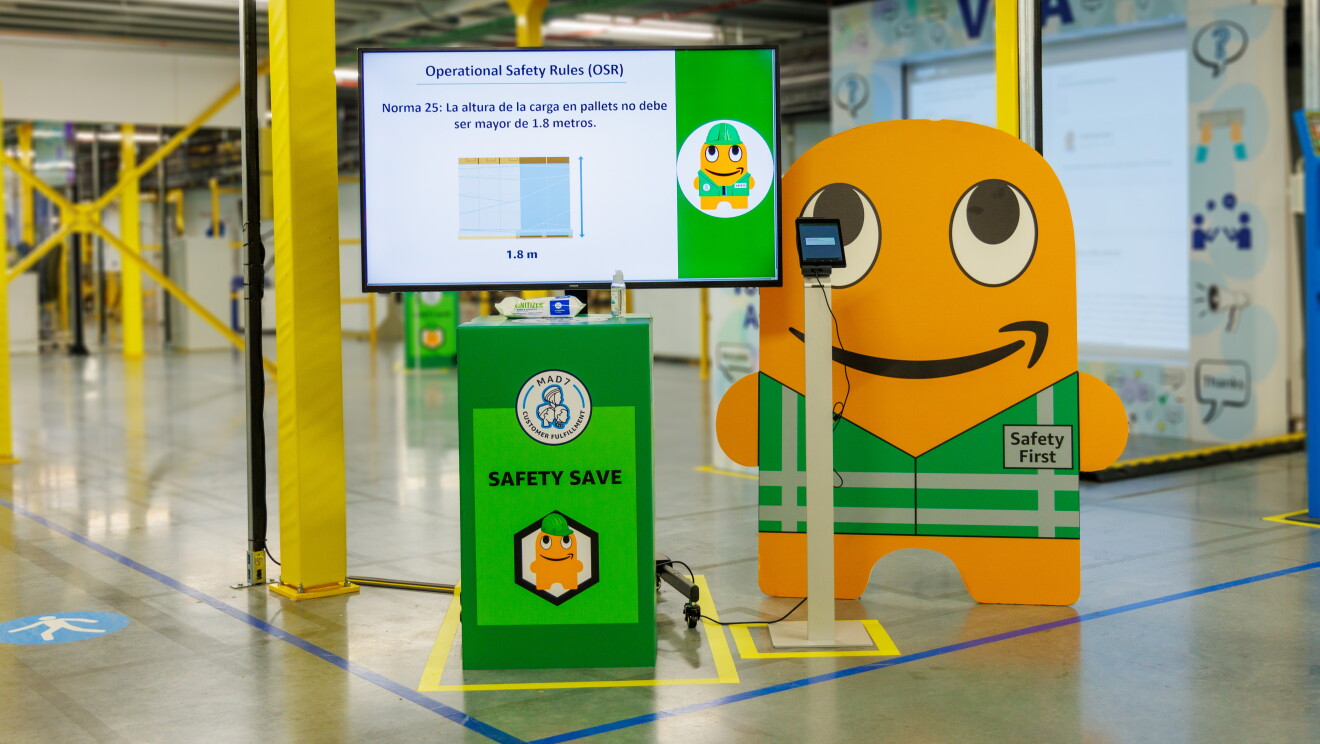 Centro logístico de Amazon. En la entrada hay una pantalla  de TV apoyada en un mostrador de color verde con gel y toallitas. Al lado una tablet con Peccy, la mascota de seguridad Peccy, detrás. Peccy es la mascota de seguridad y está en cartón, es de color naranja con la boca con la sonrisa de Amazon. Lleva un chaleco dibujado de color verde con una chapa de Safety First. Peccy también está en la pantalla de la televicsión con la Norma 25: La altura de la carga en palets no debe ser mayor de 1,8 metros. 