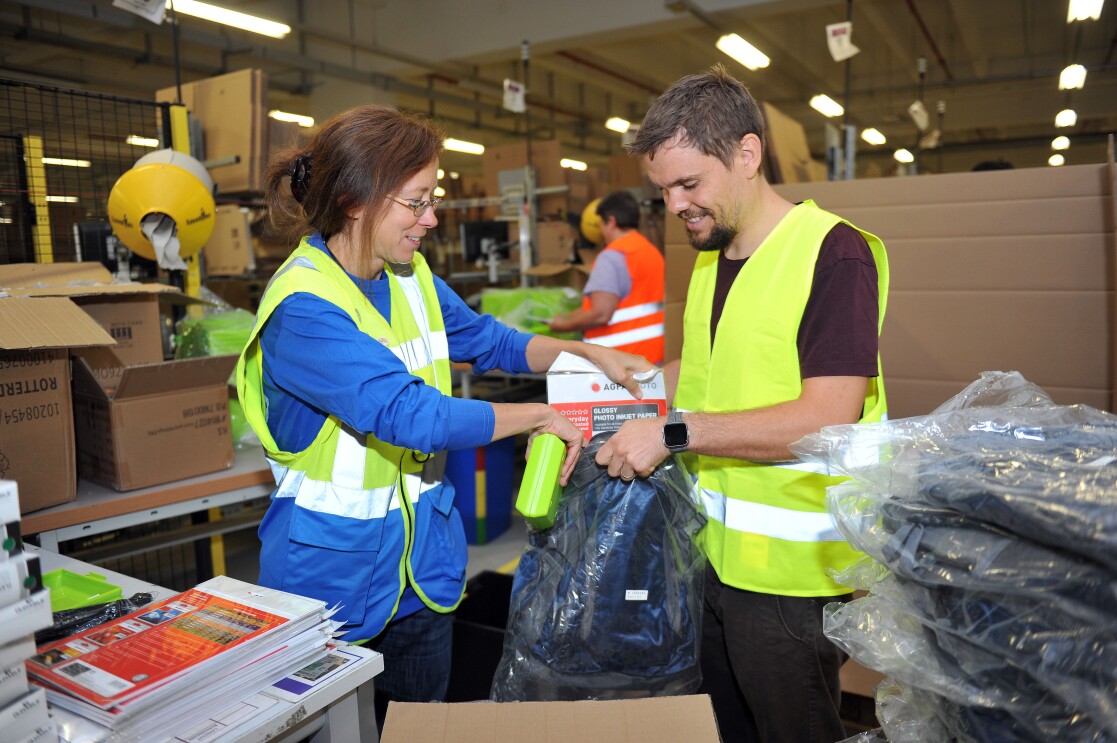 Eine Logistikmitarbeiterin in gelb-blauer Weste packt mit ihrem Kollegen aus der Verwaltung. Er trägt eine gelbe Sicherheitsweste.