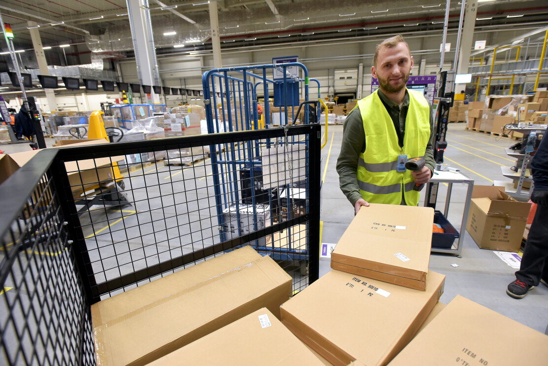 Ein Logistikmitarbeiter mit gelber Sicherheitsweste steht vor einem Packwagen. In der einen Hand hält er einen Karton, in der anderen einen Scanner.