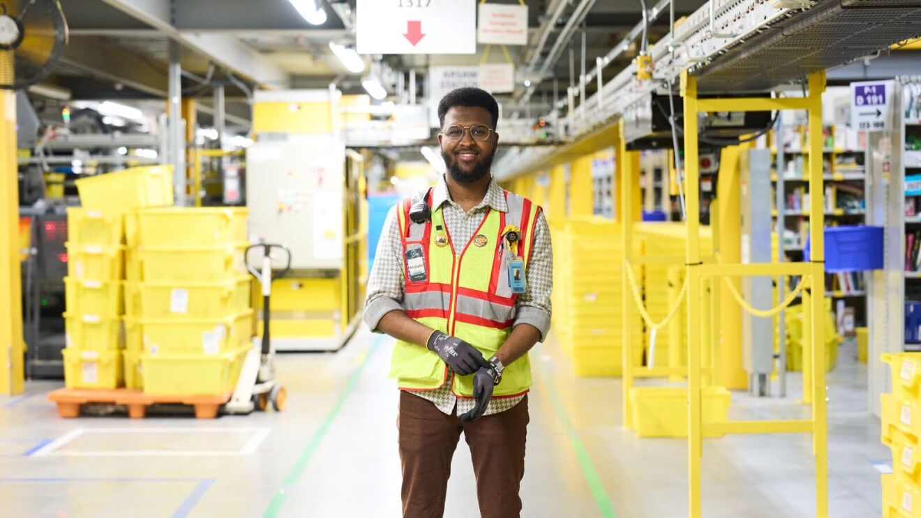 An image of Abdiaziz, an area manager working in an Amazon fulfillment center, wearing a yellow safety vest and gloves.