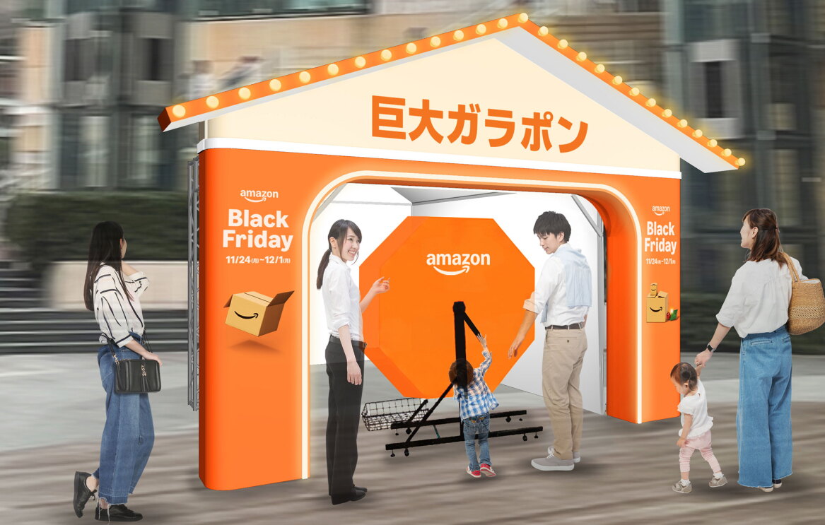 屋外に設置された、オレンジと白を基調とする抽選ブース。三角屋根状の前面に電飾と「巨大ガラポン」の文字。オレンジ色のガラポンを幼児が回している。子供の左右にスタッフと保護者、そのほかの来場者。