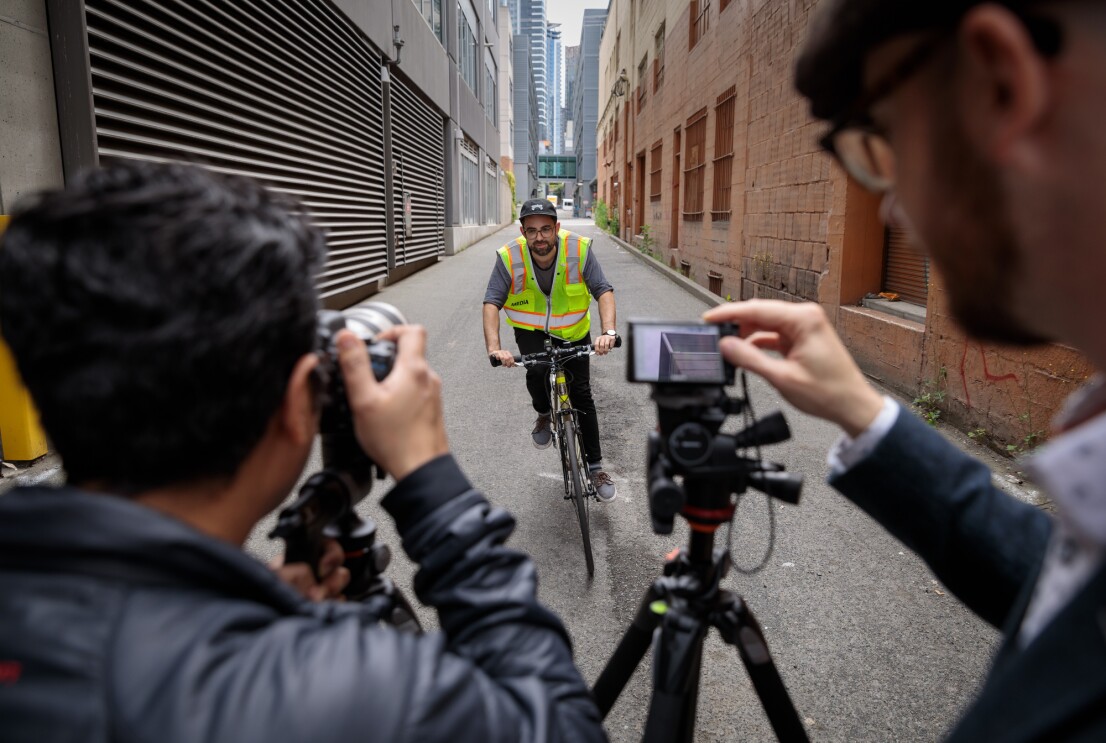 Two men in the foreground point cameras at a man on a bicycle who is riding toward them. The man on the bicycle wears a fluorescent safety vest. Buildings rise up on the right and left side of the image, and an alley stretches out behind the cyclist.