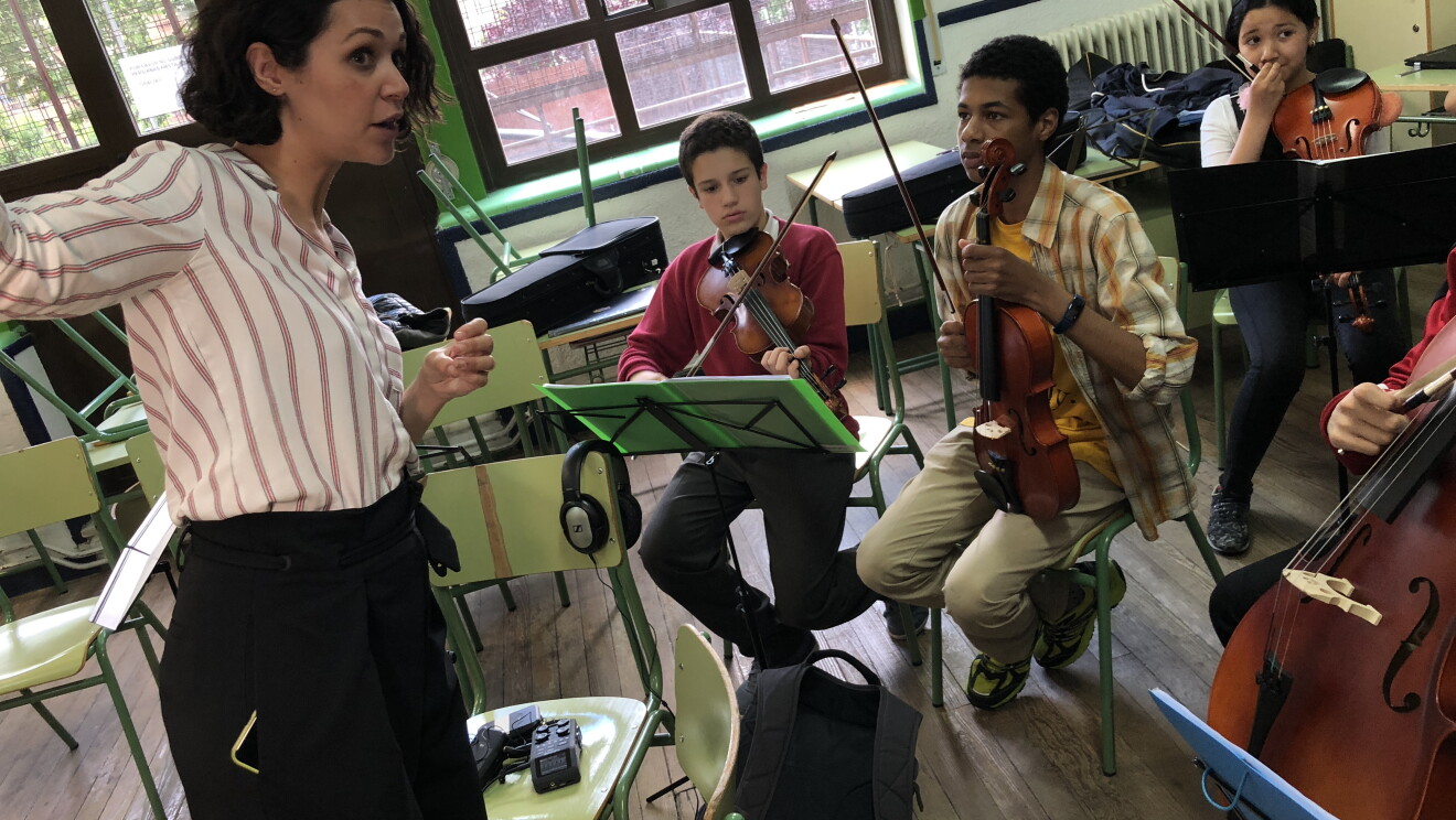 Ensayo de la camerata de Acción Social por la Música. Tres jóvenes con el violín en la mano delante de la partitura  escuchan atentamente las indicaciones de la directora de la fundación, María Guerrero. Están en una clase de instituto, las mesas y las sillas son de color verde. En el fondo, encima de las mesas, las fundas de los instrumentos. Dos grandes ventanales que dan al exterior. 