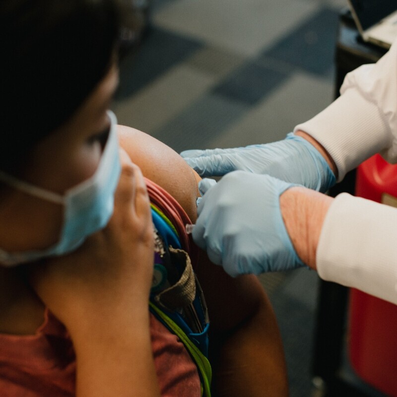 A woman wearing a mask holds up her sleeve as she receives the COVID-19 vaccine from a nurse wearing a white jacket and blue gloves.